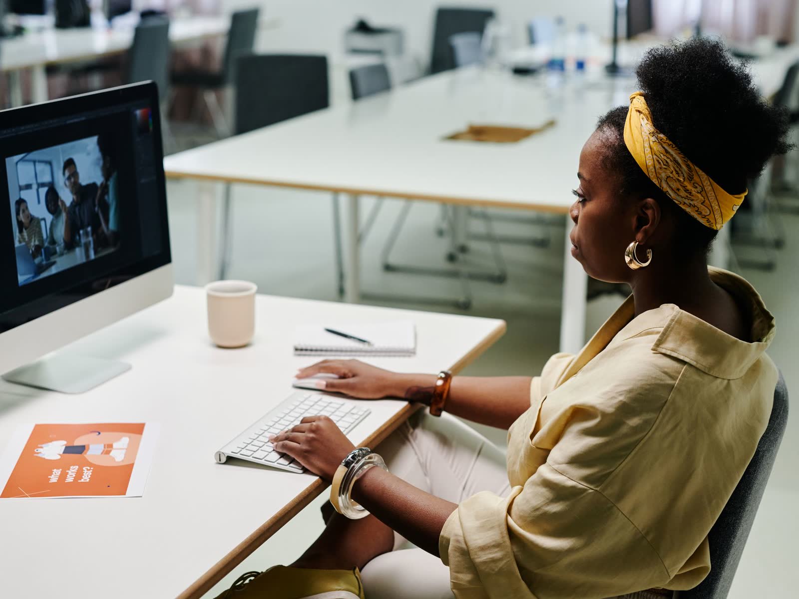 A designer working at a studio desk, editing images on a desktop computer with printed design work beside her