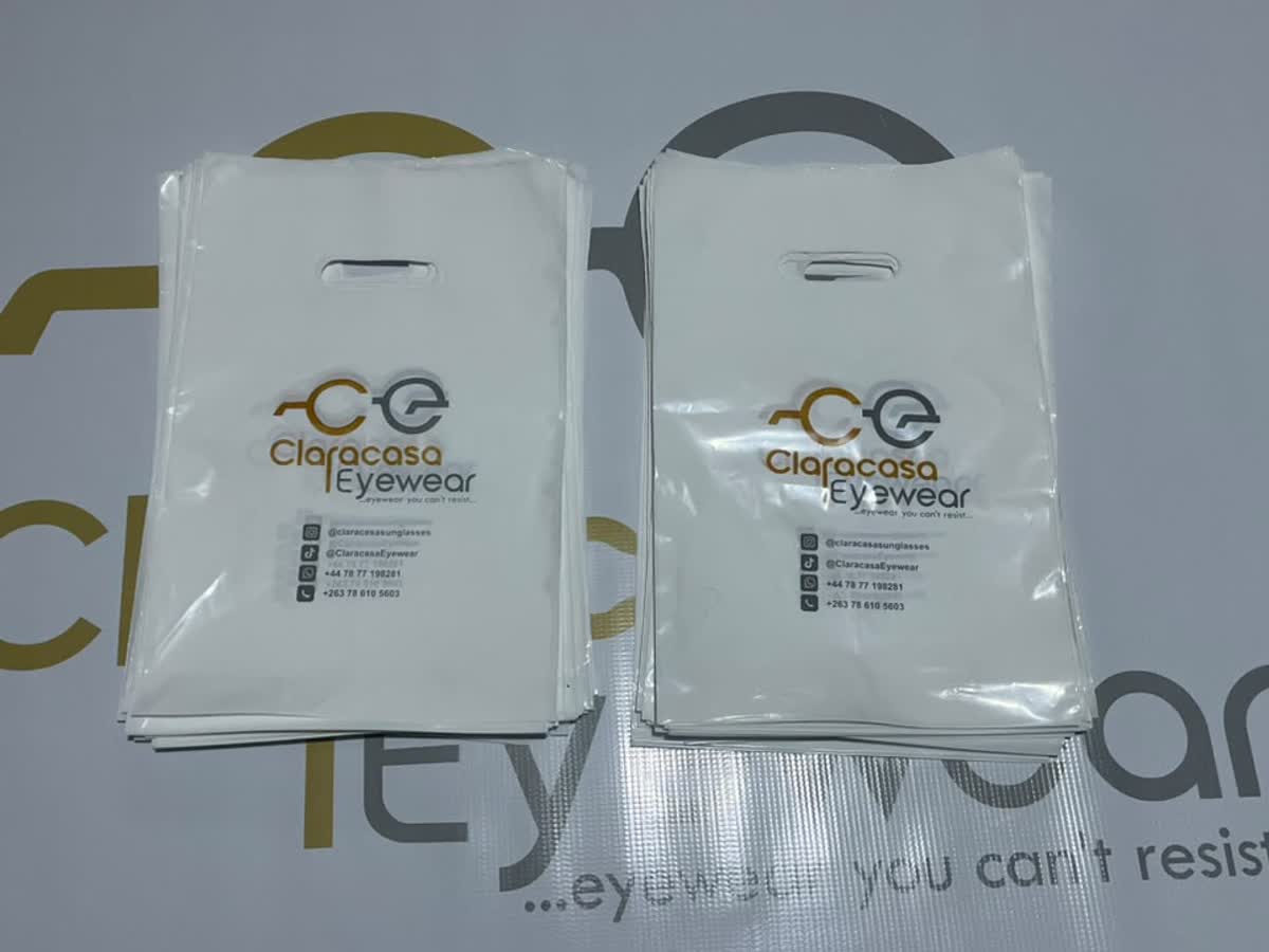 Two stacks of white Claracasa Eyewear carrier bags printed with the brand logo and contact details, photographed against a matching branded backdrop