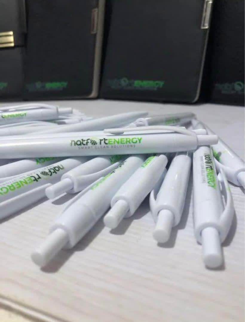 A pile of white pens printed with the Natfort Energy logo, with branded notebooks behind them