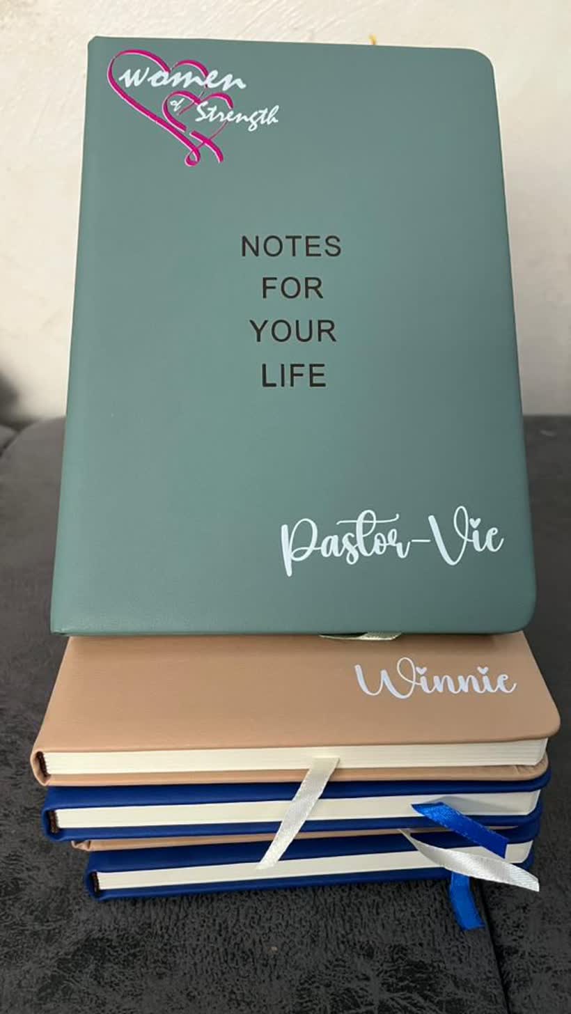 A stack of personalised notebooks, the top one teal with the Women of Strength logo and the words Notes For Your Life